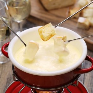 Close up of cheese fondue in a red fondue pot with 3 skewers of cubed sourdough bread dipping into the creamy, decadent cheese. Champagne in champagne glasses on table.